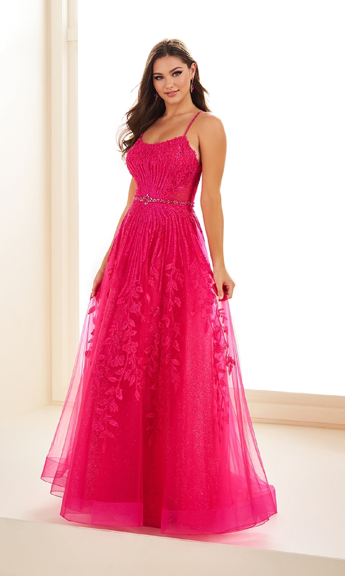 A woman stands indoors wearing the Ellie Wilde EW36068, a long prom dress in bright pink with thin straps, a beaded waistband, and floral embroidery on glitter tulle.